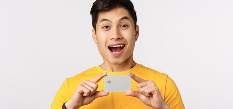 Check out this bank offer. Excited smiling and amused young asian man promote banking service, holding credit card, advice get account, paying with cashback, standing white background.