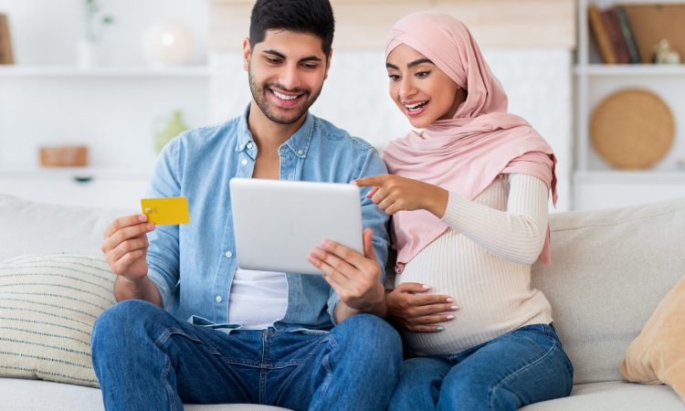 Excited pregnant islamic spouses shopping online with tablet and credit card at home while relaxing together on sofa in living room. Happy expectant arab couple buying baby clothes in internet