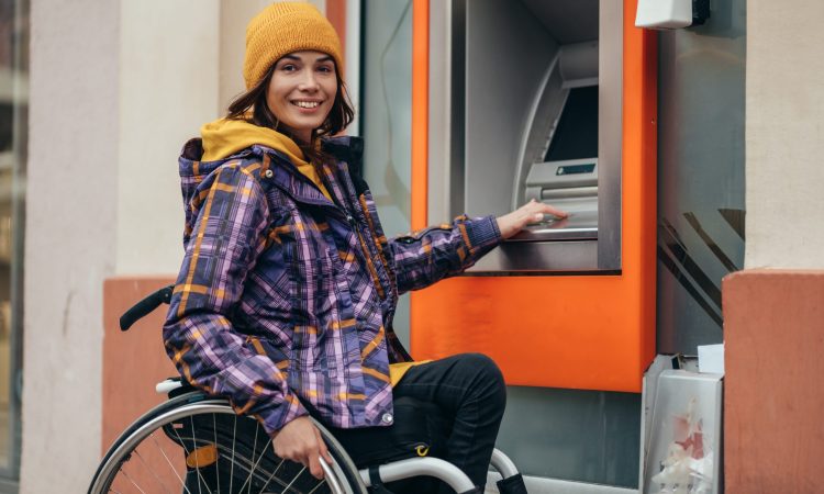 Young beautiful woman with disability who uses a wheelchair using credit card and atm machine while withdrawing money