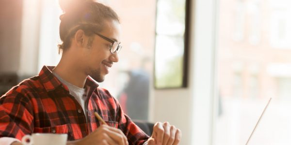 Happy young Hispanic man watching online conference and making notes in notepad