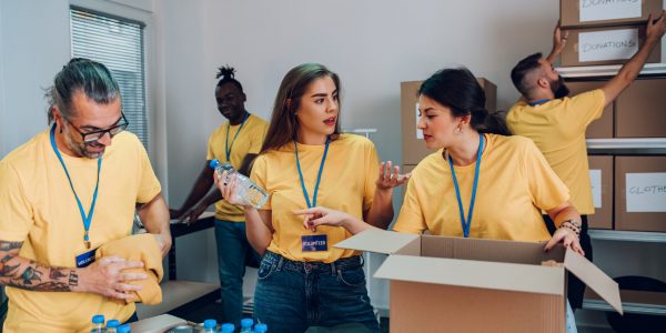 Diverse group of people working in charitable foundation. Team of multiracial volunteers in yellow uniform packing food in cardboard boxes and working together on donation project indoors. Copy space.