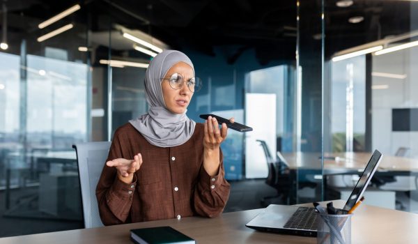 Worried young Muslim businesswoman in hijab sitting in office and talking on the phone through loudspeaker, recording voice message, solving questions, problems.