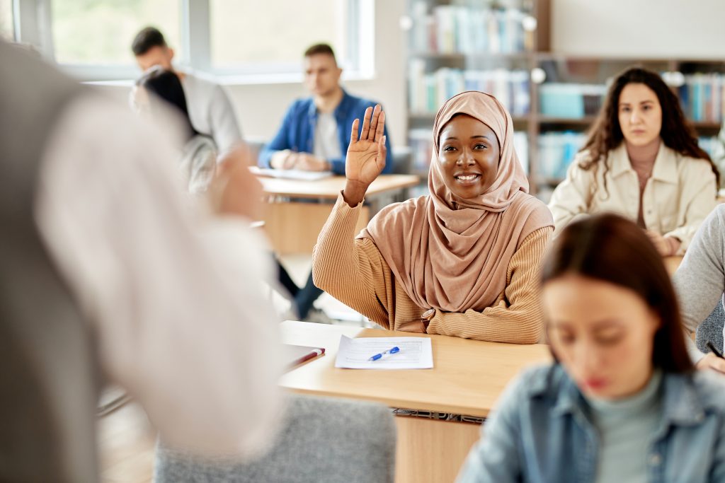 woman wearing a hijab raises her hand while seated in a classroom
